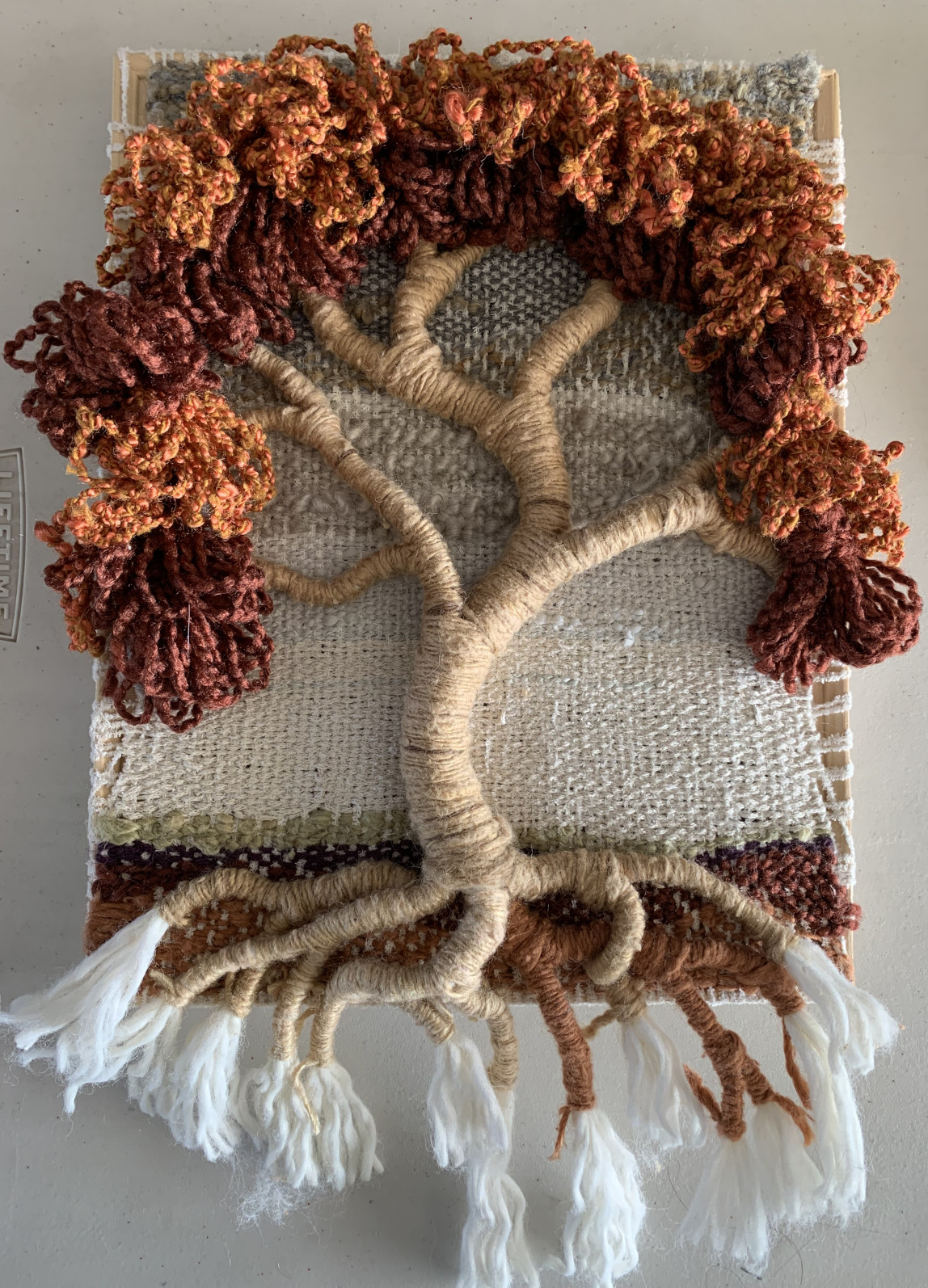 A piece of woven art with a sculptural tree; the trunk is crafted of wrapped yarn, and the leaves are draping bundles of orange and brown yarn. The roots dangle down, hanging below the wood frame.